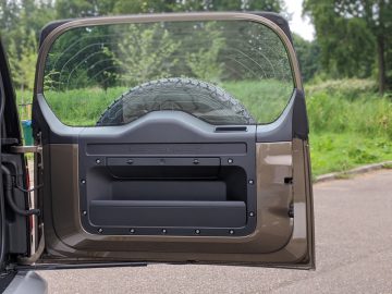 Een vrijstaande Land Rover Defender-autodeur met een gebroken raam, rustend op een weg naast een grasveld.