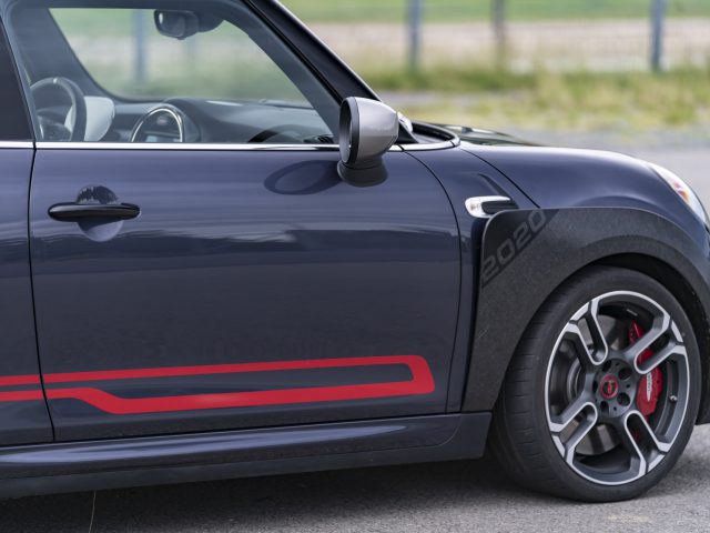 Zijaanzicht van een donkergekleurde MINI John Cooper Works GP 2020-auto met een rode accentstreep, met de nadruk op het voorwiel en de zijdeur.