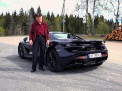 Een oudere heer staat naast een zwarte McLaren 720S-sportwagen langs de kant van de weg met een bos op de achtergrond.