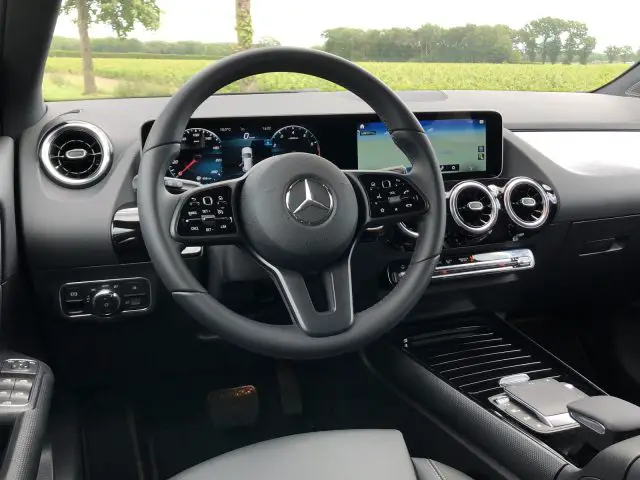 Binnenaanzicht van een Mercedes-Benz GLA-auto met een stuur, een digitaal dashboard en een infotainmentsysteem met een landelijk landschap zichtbaar buiten de ramen.