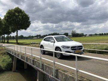 Een witte Mercedes-Benz GLA die een smalle brug over een kanaal oversteekt in een landelijke omgeving.