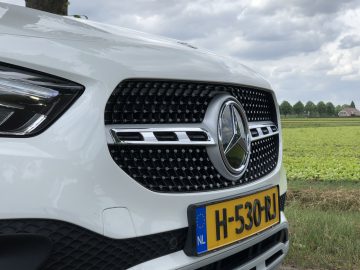 Close-up van de grille van een witte Mercedes-Benz GLA-auto en gedeeltelijk zicht op de koplamp, met een Nederlands kenteken, geparkeerd in een landelijke omgeving.