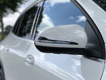 Close-up van de zijspiegel van een Mercedes-Benz GLA met een weerspiegeling van de lucht en de bomen.