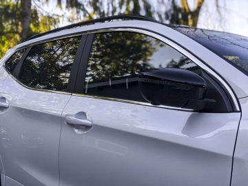 Zijaanzicht van een zilveren Nissan Qashqai N-TEC, gericht op het raam en de zijspiegel, met bomen weerspiegeld in het raam.