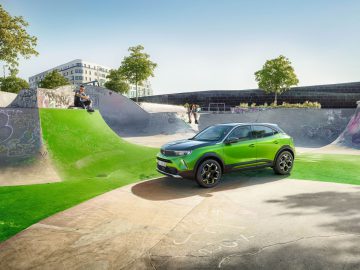 Een groene elektrische SUV van Opel Mokka geparkeerd in een met graffiti bedekt skatepark met skaters op de achtergrond.