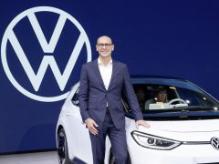 Man in zakelijke kleding staat naast een witte Volkswagen-auto op een auto-evenement, met Ralf Brandstätter in de buurt.