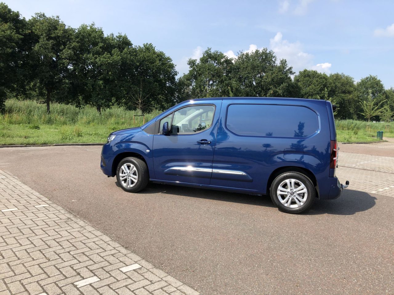 Blauwe Toyota PROACE City bestelwagen geparkeerd op een verhard terrein met gras en bomen op de achtergrond.