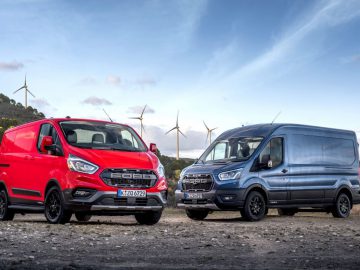 Twee Ford Transit Trail-bestelauto's staan naast elkaar geparkeerd met op de achtergrond windturbines.