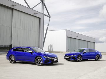 Twee blauwe Arteon sedans stonden naast elkaar geparkeerd voor een industrieel gebouw onder een bewolkte hemel.