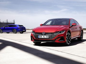 Twee Volkswagen-voertuigen, een rode Arteon op de voorgrond en een blauwe hatchback op de achtergrond, geparkeerd op een uitgestrekt asfalt onder heldere hemel.