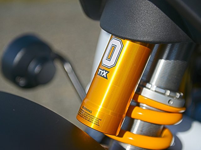 Close-up van een geel-zilveren Ariel Nomad R-schokdemper met het Ohlins-merklogo, opgenomen met een geringe scherptediepte.