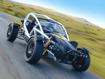 Een Ariel Nomad R-buggy met rolkooi die over een open weg rijdt, met een helderblauwe lucht en met gras begroeide heuvels op de achtergrond.