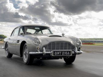 Een klassieke zilveren Aston Martin DB5 Goldfinger Continuation rijdend op een asfalt, met wolkenluchten op de achtergrond.