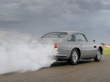 Een vintage Aston Martin DB5 accelereert op een circuit en stoot van achteren een wolk witte rook uit, onder een bewolkte hemel.