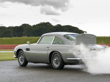 Een zilveren Aston Martin DB5 Goldfinger Continuation die van achteren rook uitstraalt op een racecircuit, met een met gras begroeide en bewolkte achtergrond.