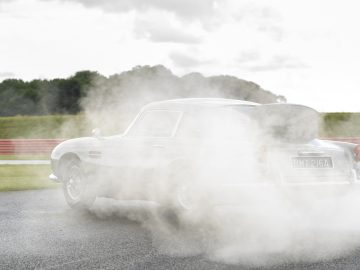 Een Aston Martin DB5 Goldfinger Continuation slipt op een racecircuit, omringd door een rookwolk.