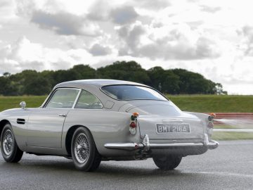 Een vintage zilveren Aston Martin DB5 Goldfinger Voortzetting rijdend op een weg met een landelijk landschap op de achtergrond.