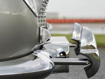 Close-up van de chromen bumper en koplampen van een Aston Martin DB5 Goldfinger Continuation, met een wazige racebaanachtergrond.