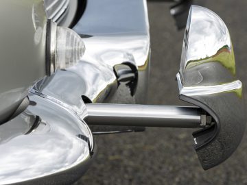 Close-up van de zijkant van een Aston Martin DB5 Goldfinger Continuation met chromen bumpers en koplamp met een opvallend ontworpen zijspiegel.