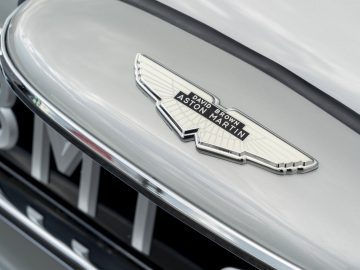 Close-up van een Aston Martin DB5 Goldfinger Continuation-logo op de motorkap van een zilveren auto.