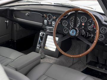 Binnenaanzicht van een Aston Martin DB5 Goldfinger Continuation met een houten stuur, witleren stoelen en een gedetailleerd dashboard met vintage meters.