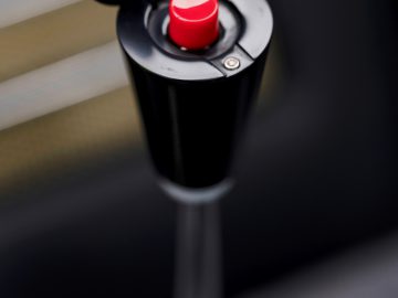 Close-up van een noodstopknop op een machine, gemarkeerd met selectieve focus, tegen een wazige Aston Martin DB5 Goldfinger Continuation-achtergrond.