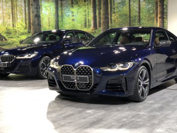 Twee BMW-auto's, een blauwe 4 Serie Coupé en een grijze 5 Serie, tentoongesteld in een overdekte showroom met een bosachtergrond.
