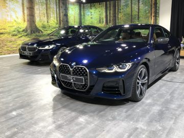 Blauwe BMW 4 Serie Coupé tentoongesteld in een showroom met een ander BMW-model op de achtergrond, tegen een achtergrond met bosthema.