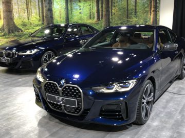 Twee BMW 4 Serie Coupé-auto's tentoongesteld tijdens een indoorevenement, één een blauwe coupé op de voorgrond en een zwarte sedan op de achtergrond, beide tegen een bosachtergrond.