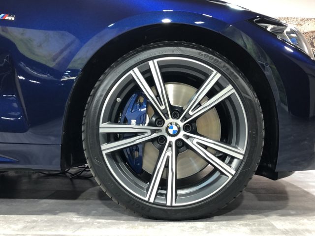 Close-up van een BMW 4 Serie Coupé-wiel met het blauwe BMW-logo op de wieldop en geïnstalleerd op een blauwe auto, binnenshuis weergegeven.