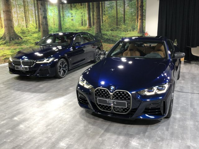 Twee BMW-auto's tentoongesteld op een binnententoonstelling, de ene een blauwe BMW 4 Serie Coupé en de andere een grijze stationwagen, beide tegen een achtergrond met bosthema.