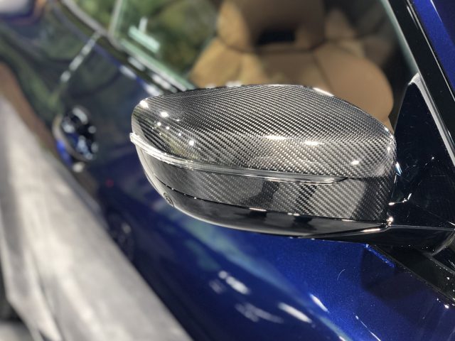 Close-up van de zijspiegel van een BMW 4 Serie Coupé met een koolstofvezelkap op een blauw voertuig.