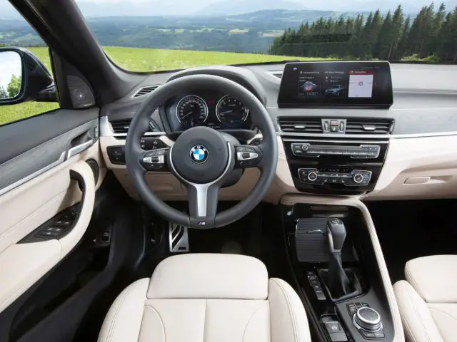 Binnenaanzicht van een BMW X1 met het stuur, het dashboard en de leren stoelen, terwijl een schilderachtig landschap zichtbaar is door de voorruit.