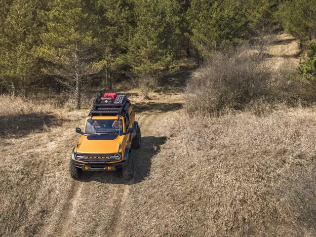 Luchtfoto van een gele Ford Bronco die over een onverharde weg door een dun bos rijdt.