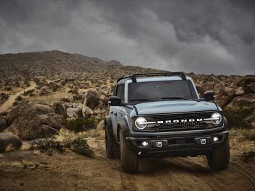 Een blauwe Ford Bronco die onder een bewolkte hemel over een ruige onverharde weg rijdt in een rotsachtig woestijnlandschap.