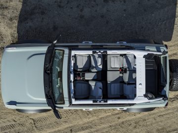 Luchtfoto van een lichtblauwe Ford Bronco met een open bovenkant, waardoor een zitopstelling met drie rijen zichtbaar is, geparkeerd op een zanderige ondergrond.