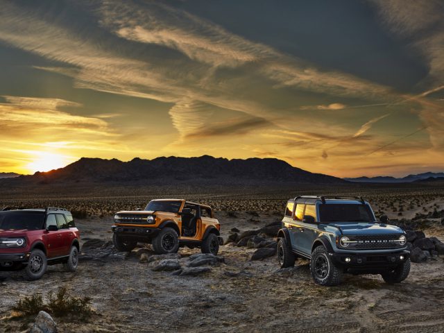 Drie Ford Bronco's geparkeerd in een woestijn bij zonsondergang met bergen op de achtergrond en een levendige lucht gemarkeerd door contrails.