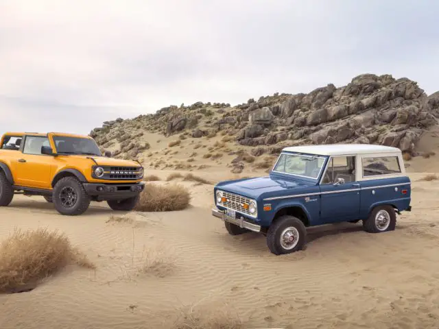 Twee SUV's, een moderne Ford Bronco in oranje en een vintage in blauw, geparkeerd in een zandwoestijnlandschap met rotspartijen op de achtergrond.