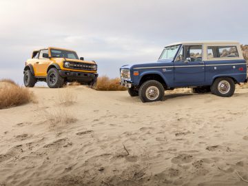 Twee terreinwagens, een moderne gele Ford Bronco en een klassieke blauwe SUV, geparkeerd op een zandwoestijnheuvel met rotsachtige achtergrond.