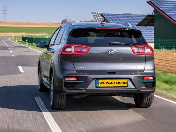 Een grijze Kia Cee'd, een EV met overheidssubsidies, rijdend op een weg met zonnepanelen en elektriciteitsleidingen op de achtergrond.