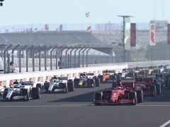 Formule 1-auto's opgesteld op een racecircuit bij de startgrid, met een heldere hemel en toeschouwerstribunes zichtbaar op de achtergrond tijdens het F1 2020-seizoen.