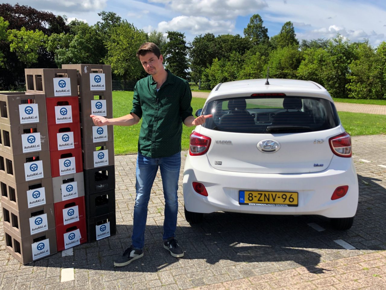 Een man staat tussen een stapel producten in dozen en een witte Hyundai i10 hatchback in een zonnige parkomgeving.