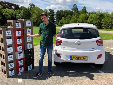 Een man staat tussen een stapel producten in dozen en een witte Hyundai i10 hatchback in een zonnige parkomgeving.