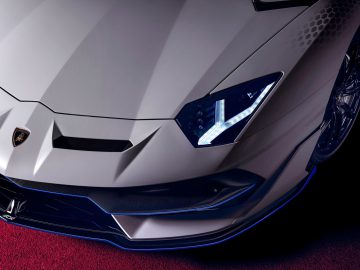 Close-up van een zilveren Lamborghini Aventador SVJ, gefocust op de motorkap en koplamp, met een rode achtergrond.