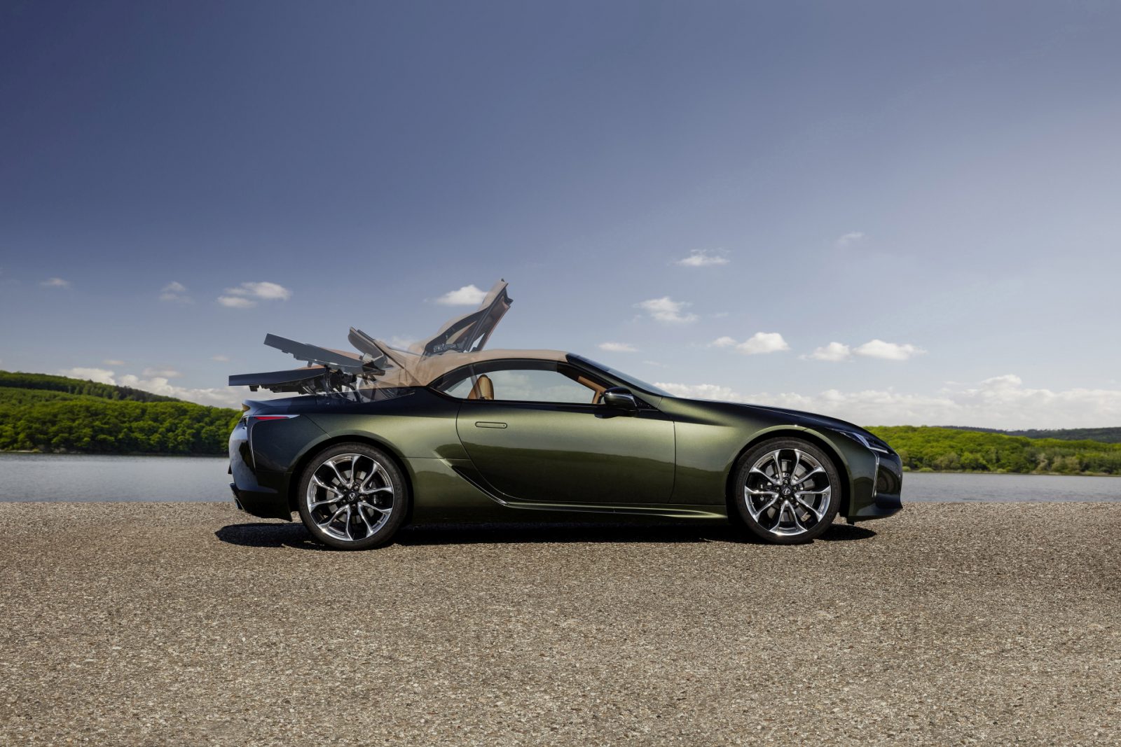 Een groene Lexus LC Convertible met de deuren naar boven open, geparkeerd aan de oever van een meer onder een helderblauwe lucht.