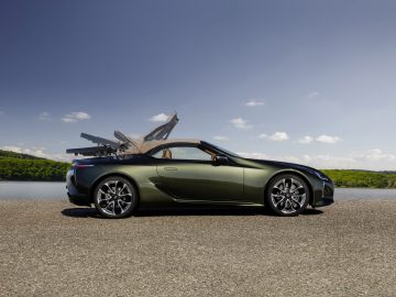 Een groene Lexus LC Convertible met de deuren naar boven open, geparkeerd aan de oever van een meer onder een helderblauwe lucht.