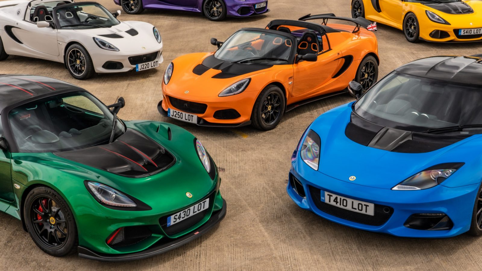 Een verzameling van vijf kleurrijke Lotus-sportwagens geparkeerd op beton, met verschillende modellen in wit, oranje, groen, blauw en geel.