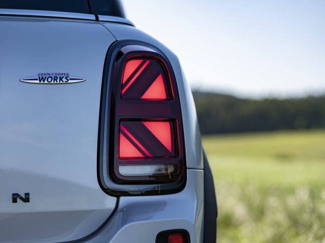 Close-up van het achterlicht en het logo aan de achterkant van een MINI John Cooper Works Countryman-model, geparkeerd met een wazig groen veld en een heldere lucht op de achtergrond.