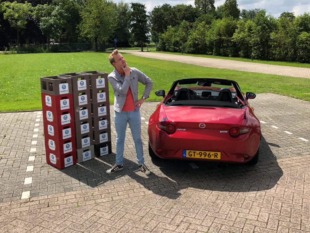 Man die naast een stapel schoenendozen staat en een telefoongesprek voert, met een rode Mazda MX5 ND-cabriolet vlakbij geparkeerd in een zonnige buitenomgeving.