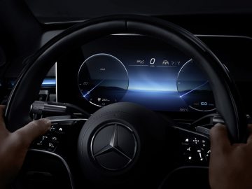 Handen van de bestuurder op het stuur van een Mercedes-Benz S-Klasse met uitzicht op digitale dashboarddisplays die snelheid en navigatie weergeven.